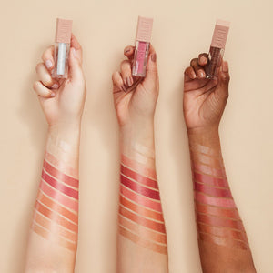 Three hands holding lip gloss applicators with color swatches on a beige background