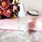 Skincare serum bottle labeled 'The Ordinary' on a fluffy surface with flowers and false eyelashes in the background.